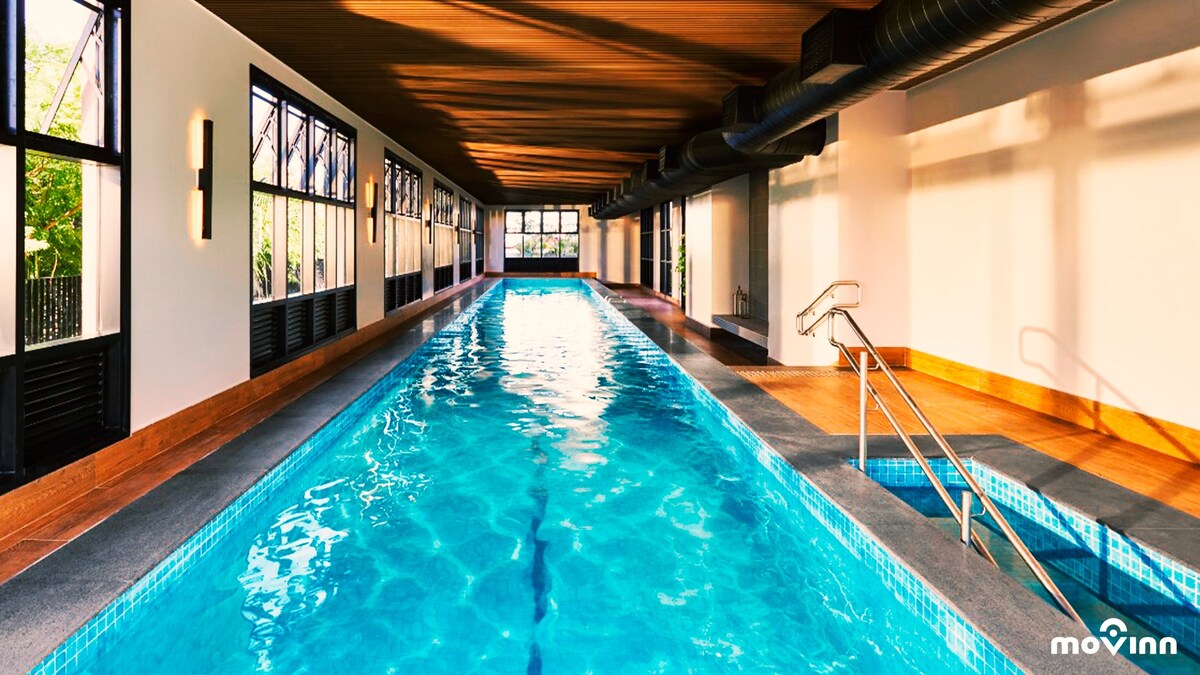 An indoor swimming pool features crystal clear water, with steps leading into it. Natural light filters through large windows along one side, illuminating the wooden ceiling and sleek tilework throughout the space. The atmosphere is designed to promote relaxation and comfort.