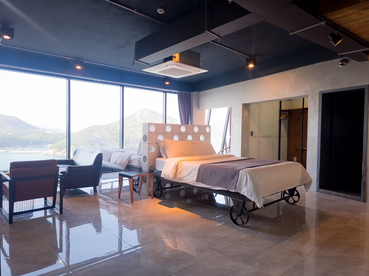 A bright room features a spacious bed with a minimalist design, accompanied by a comfortable seating area including chairs and a table. Large windows provide expansive views of the mountains and ocean, while polished tiles enhance the room's modern look.