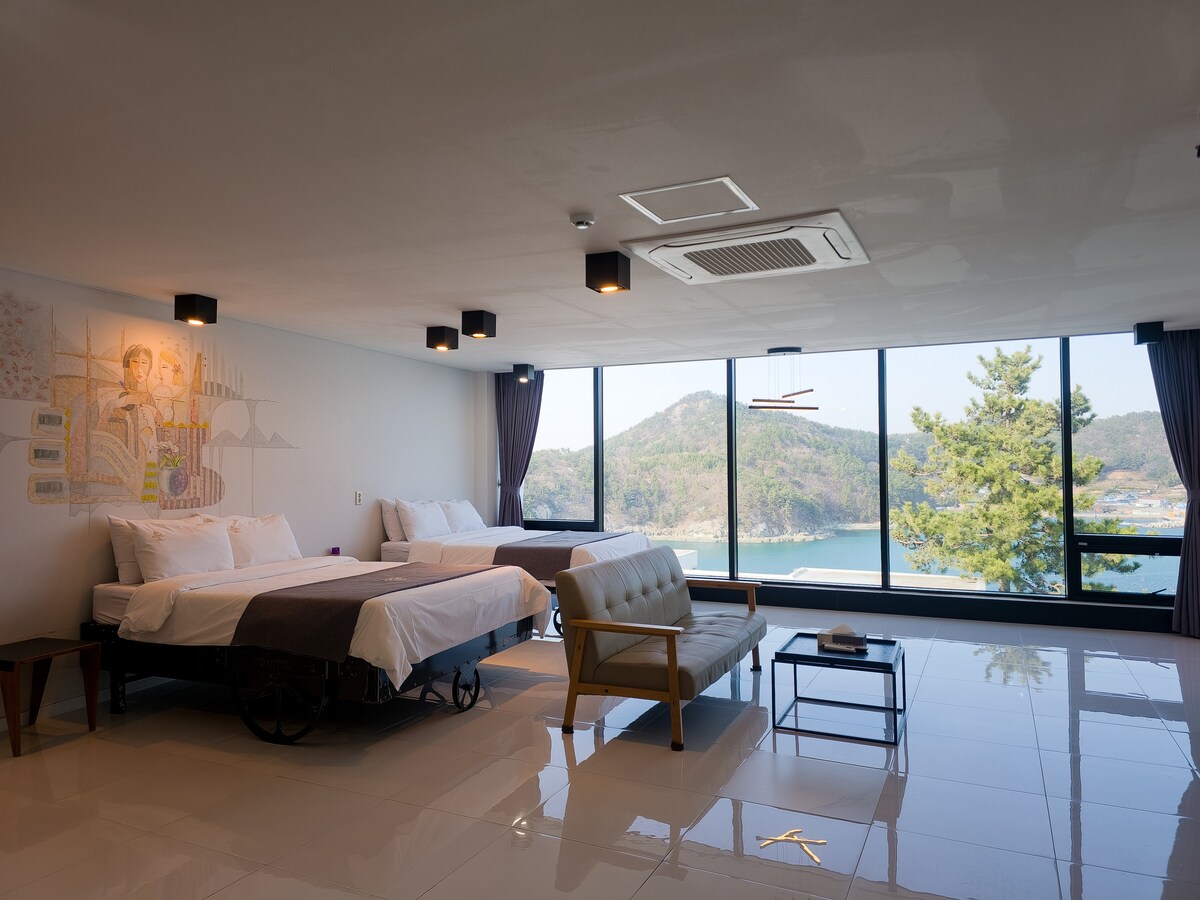 A spacious room features large windows with a view of the surrounding landscape. Two beds are positioned alongside a sleek couch and a small table. Artwork is displayed on the wall, adding a touch of creativity to the serene atmosphere.