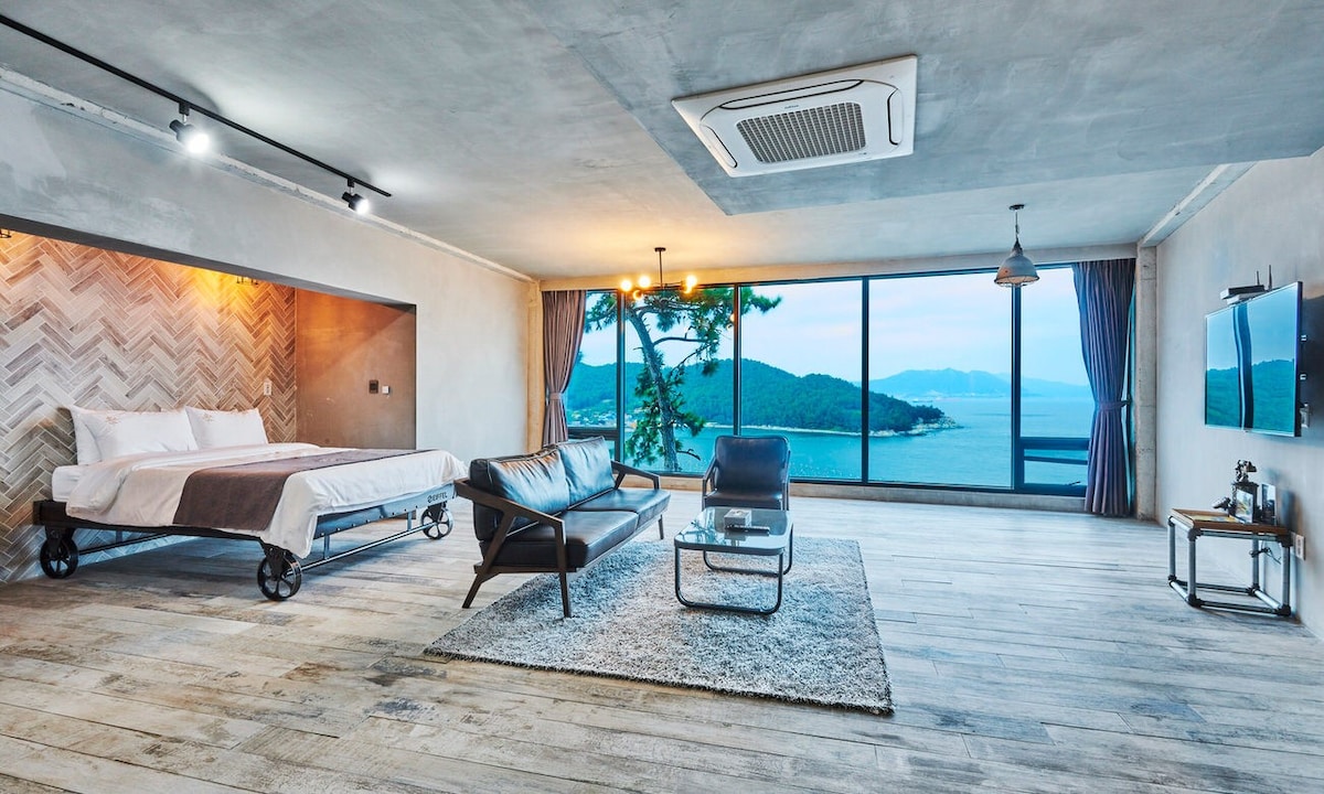 A spacious room features a large glass window revealing a stunning sea view. A comfortable bed, a sleek black sofa, and a glass coffee table are arranged on a light wooden floor. Soft lighting adds a cozy ambiance, and modern decor is visible on the walls.