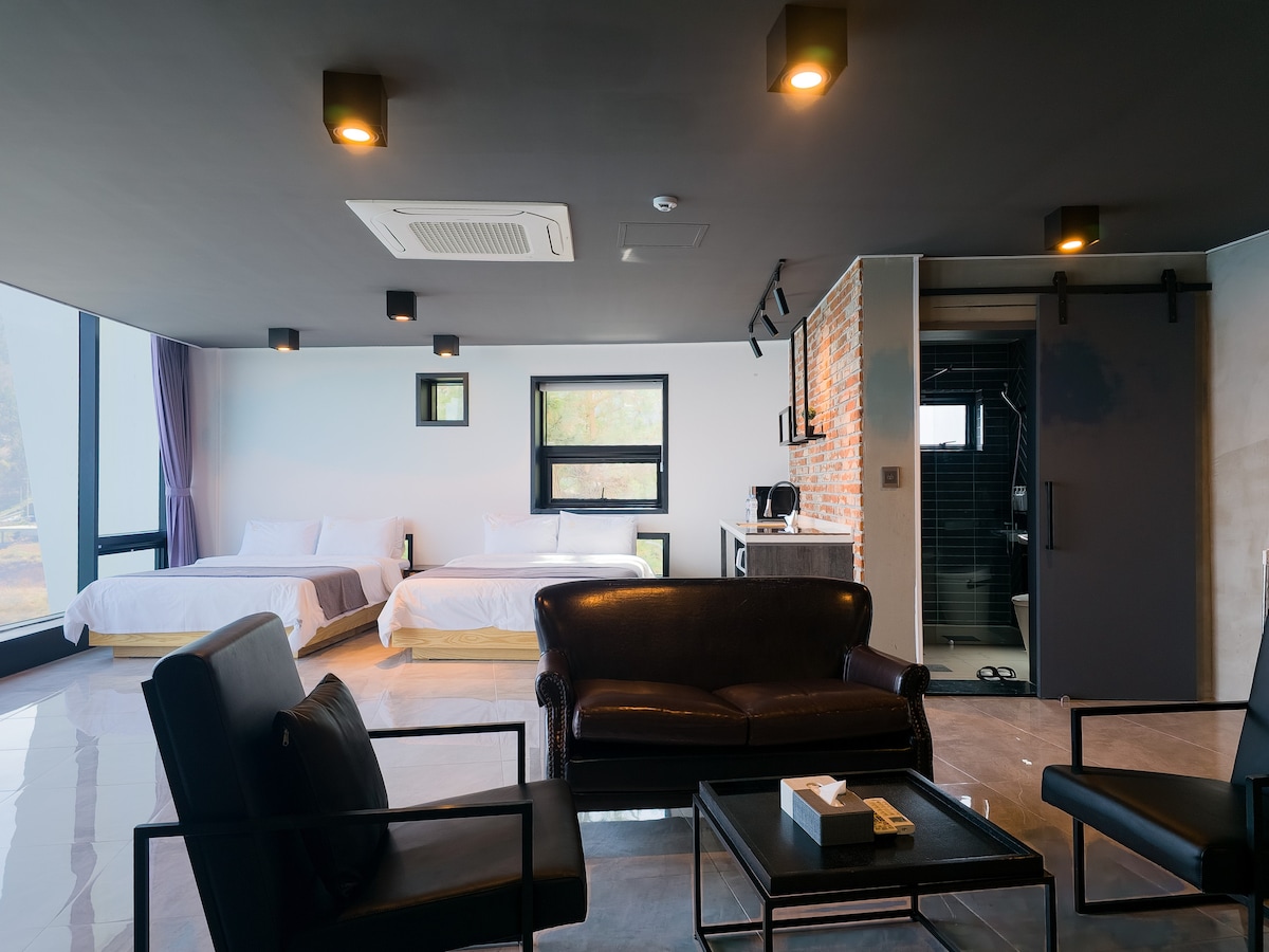 A spacious room is presented with large windows allowing natural light to illuminate the interior. Two beds are positioned near the windows, while a black sofa and chairs create a cozy seating area. A small table is centrally placed, complemented by a kitchenette and bathroom area.