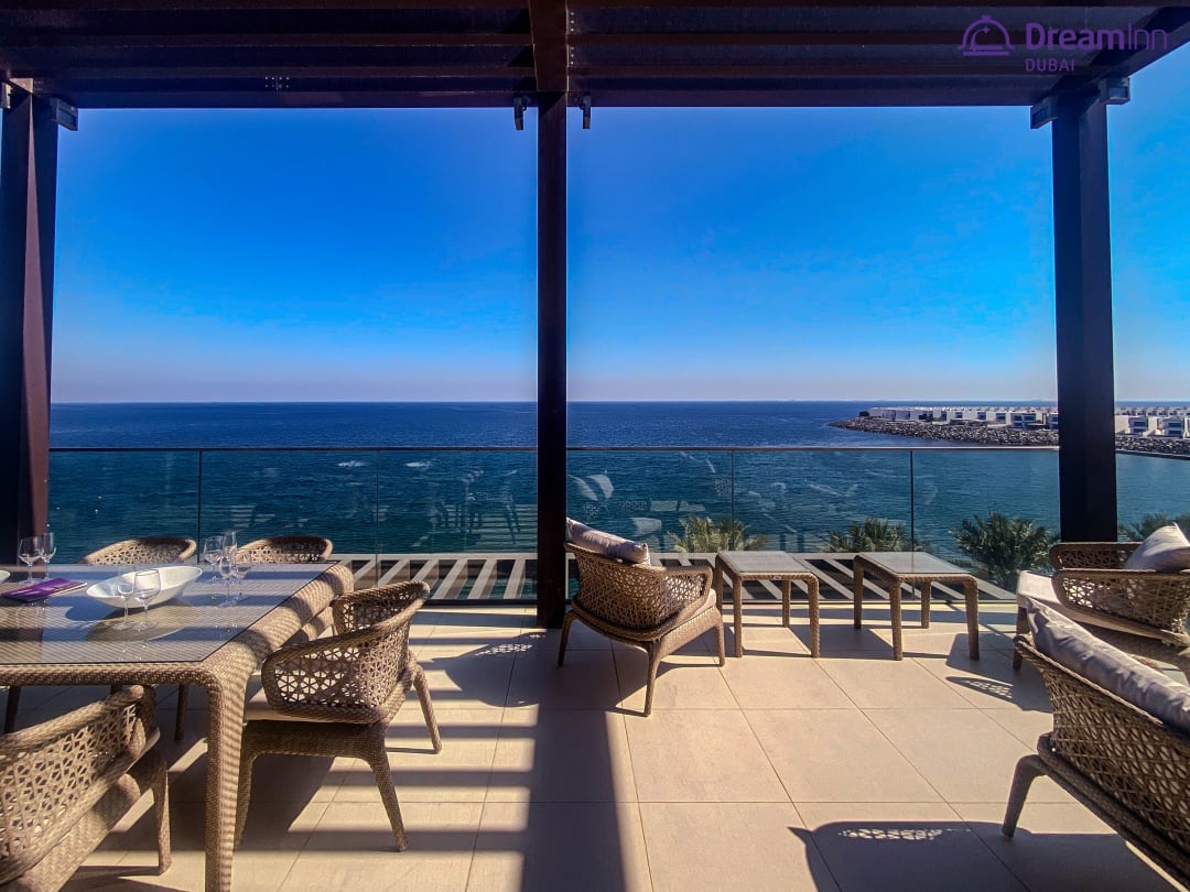 A spacious outdoor terrace is featured, overlooking a clear blue sea and sky. Comfortable seating arrangements and a dining table are provided, conducive for relaxation and enjoying meals. The area is shaded by a wooden pergola, creating a serene environment for outdoor gatherings.
