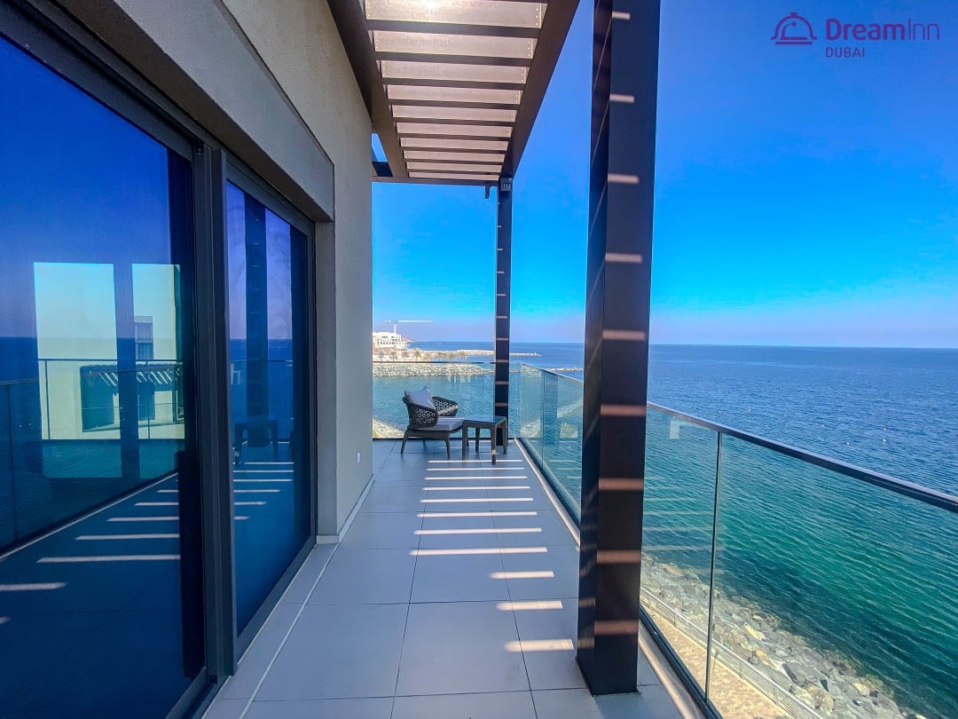 A private balcony features sleek glass railings offering views of the sea and coastline. Modern outdoor furniture is arranged against the backdrop of clear blue skies, providing a serene space for relaxation. Natural light floods the area, enhancing the inviting atmosphere.