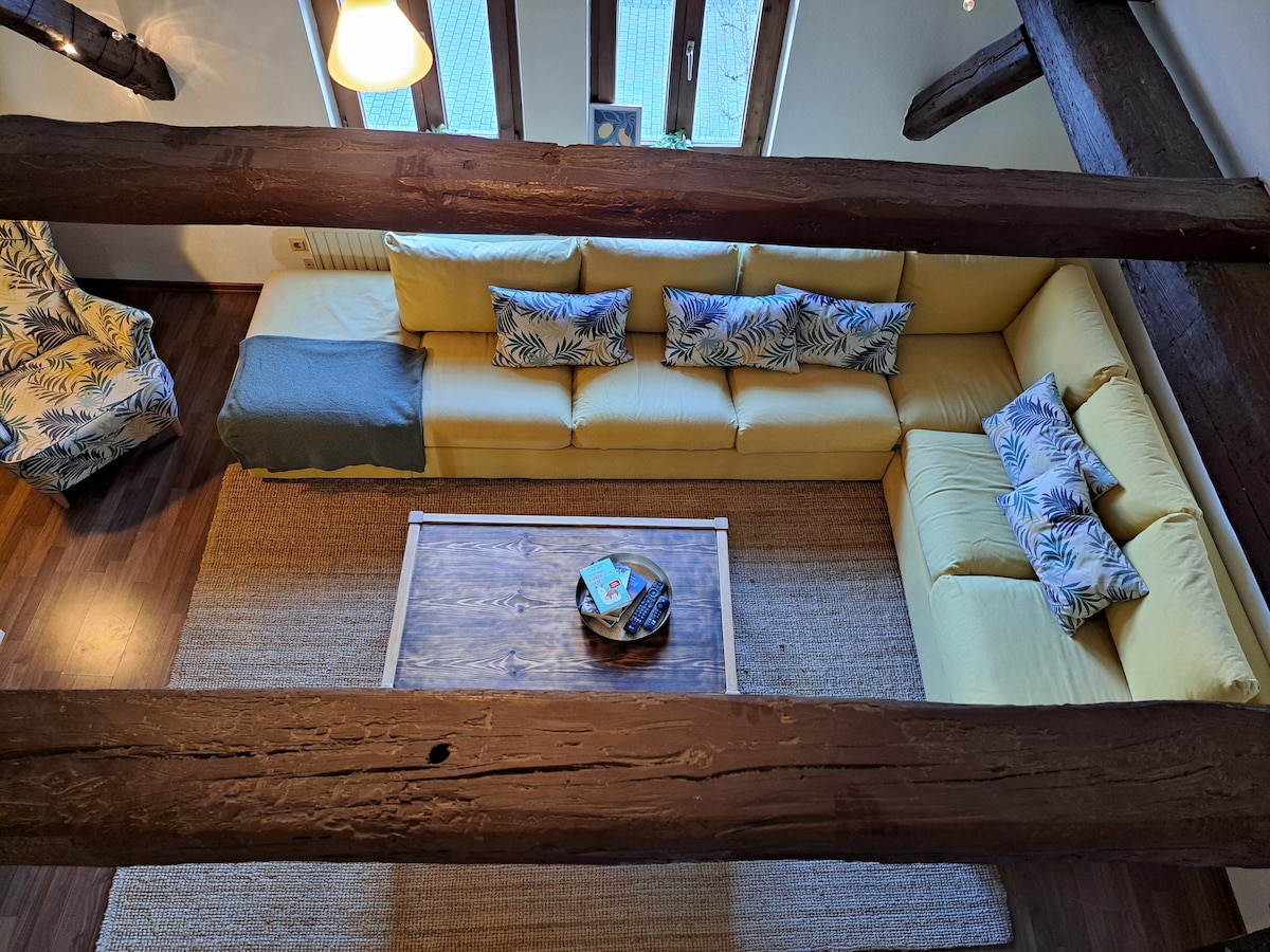 An aerial view of the living room highlights a spacious yellow sectional sofa adorned with cushions featuring a tropical print. A wooden coffee table sits centrally on a woven area rug, while exposed wooden beams add a rustic touch to the space.