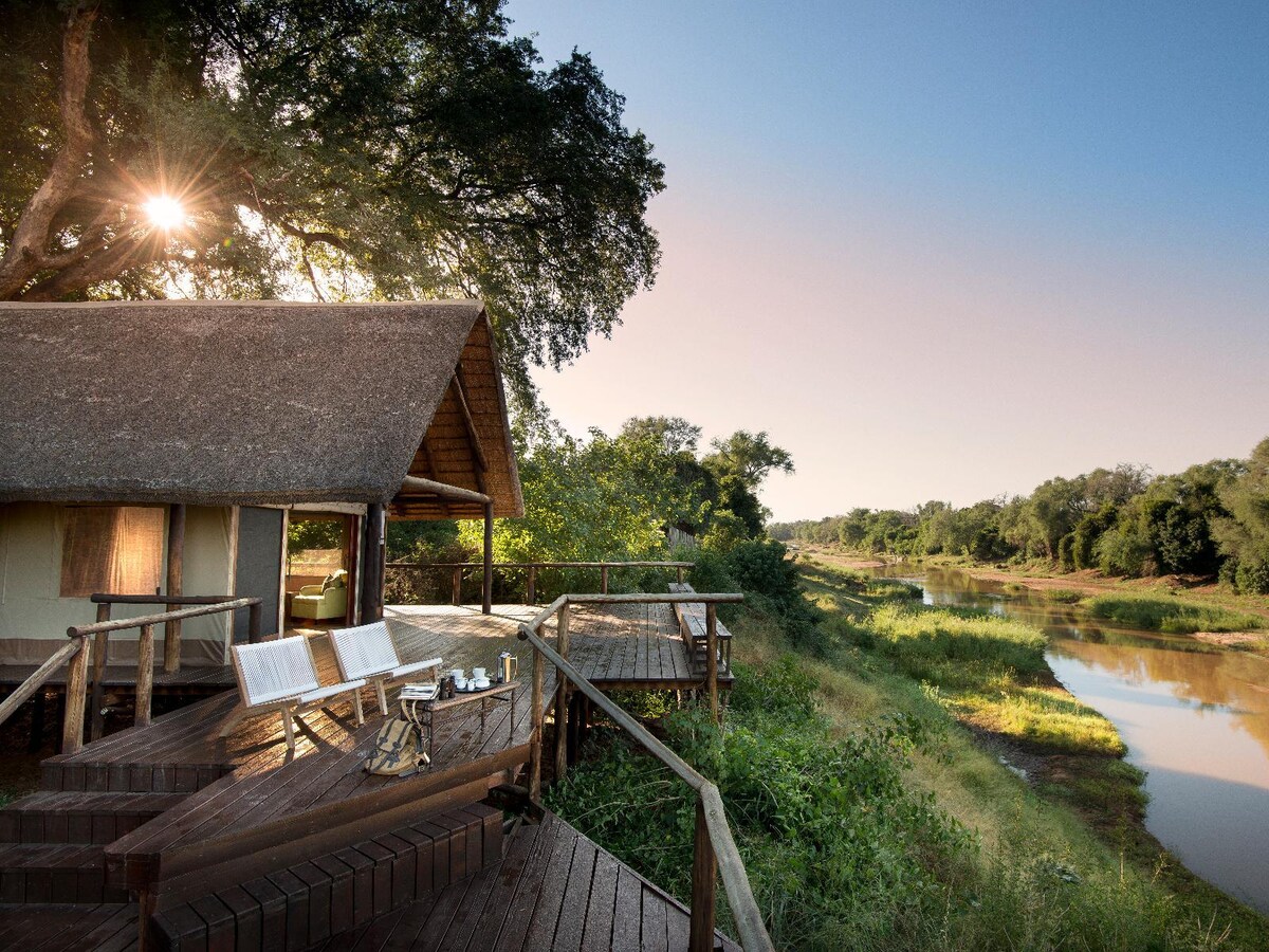 A luxury tent is positioned on a wooden deck, offering views of a serene river lined with lush greenery. Morning sunlight filters through the leaves of nearby trees, casting soft shadows on the deck, which is furnished with chairs and a small table.