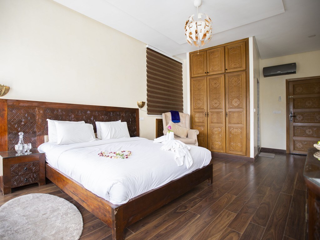 A spacious bedroom features a large wooden bed adorned with white bedding and decorative accents. A comfortable chair is positioned nearby, while a stylish area rug adds warmth to the wooden floors. Ample natural light filters through the window with brown shades, and wooden cabinetry provides storage.