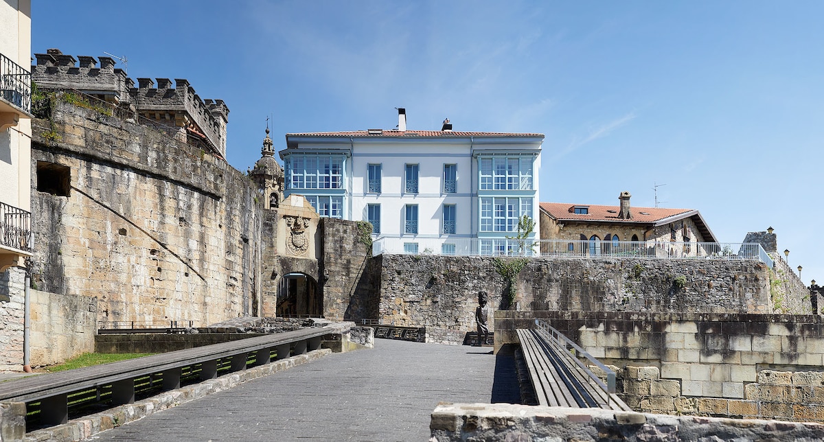 A historic stone wall frames the modern building, showcasing large windows with a blue hue. The walkway extends in front, lined with stone borders, leading towards the vibrant greenery in the background. A clear blue sky completes the outdoor scene.