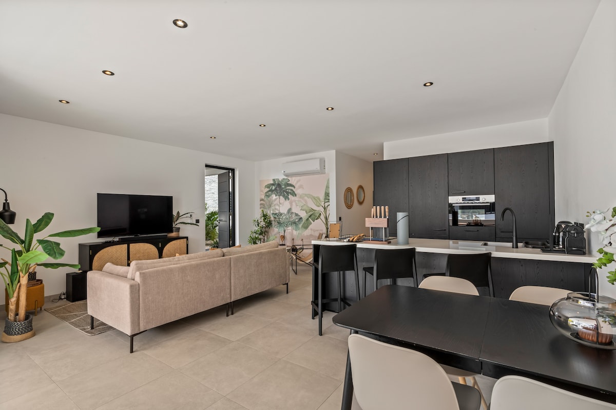 A spacious living area features a comfortable sofa set, a large smart TV mounted on the wall, and an open kitchen with modern appliances. The dining area includes a black table with matching chairs, while potted plants add a touch of greenery to the environment.