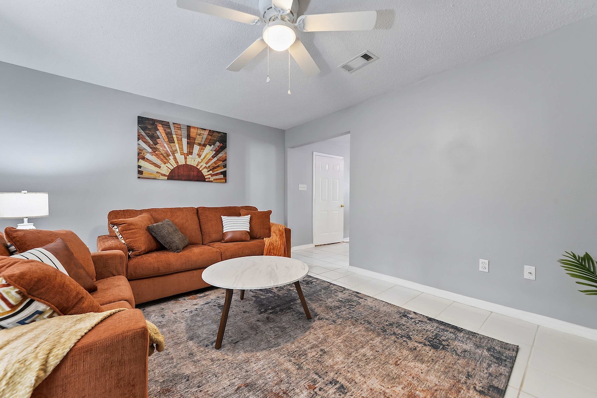 The living room features a cozy arrangement with an orange sectional sofa and a round marble coffee table. A large rug adds warmth to the space. Natural light is diffused through the doorway, and a ceiling fan provides a cool breeze, enhancing comfort.