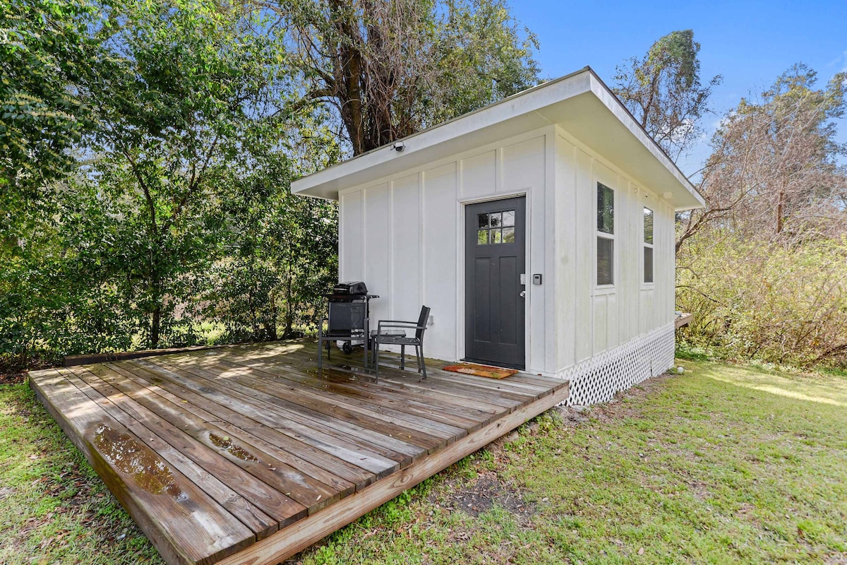 The exterior view of the Coastal Cabana highlights a modern studio with a light-colored facade set against lush greenery. A spacious wooden deck features a gas BBQ grill and a chair, providing a comfortable outdoor space. The private entrance is clearly visible.