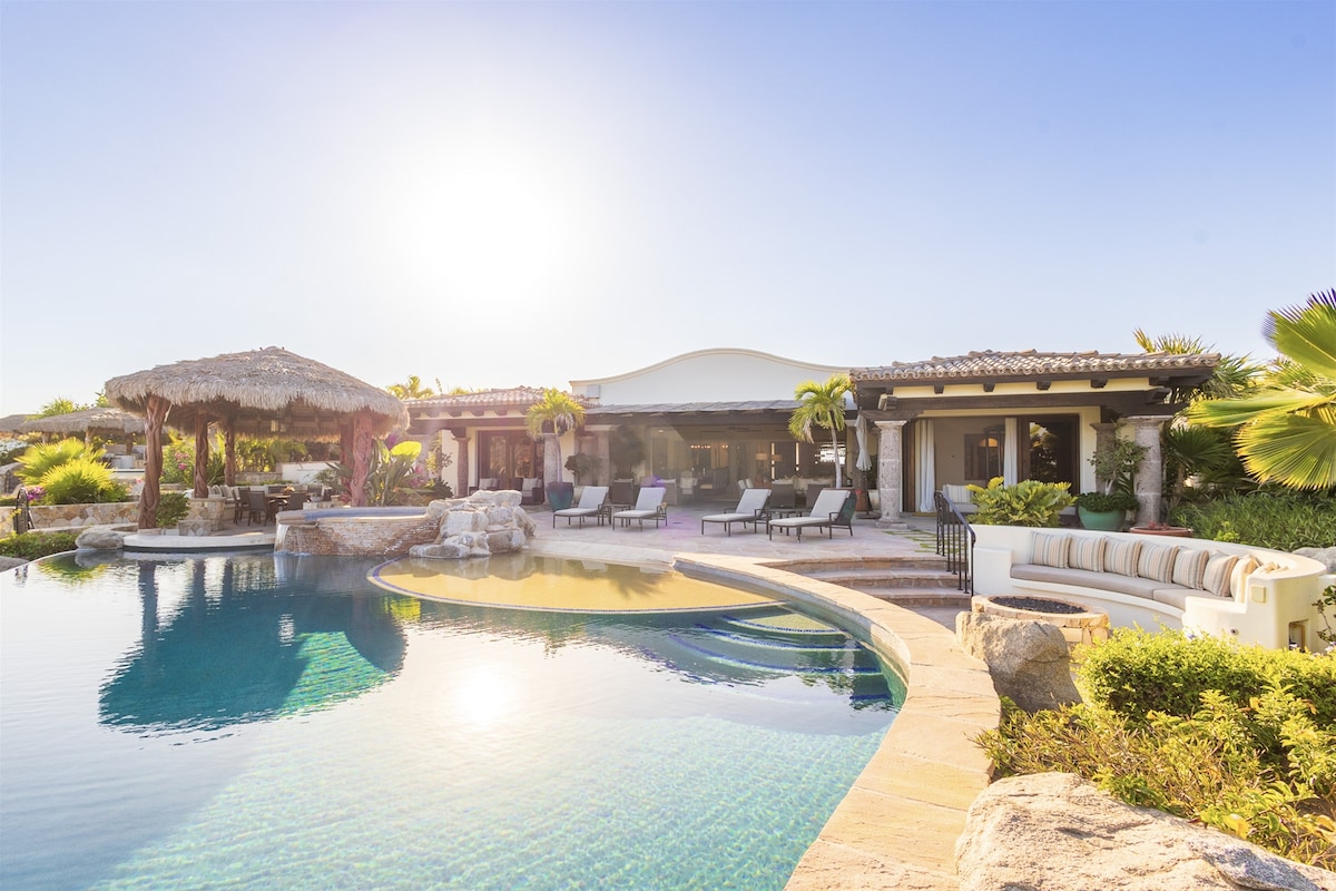 An outdoor view of a luxurious villa showcases a sparkling infinity pool with a gentle curve. A thatched roof palapa and lounge chairs are positioned nearby, surrounded by lush greenery. The sun casts a warm glow over the scene, enhancing the inviting atmosphere.