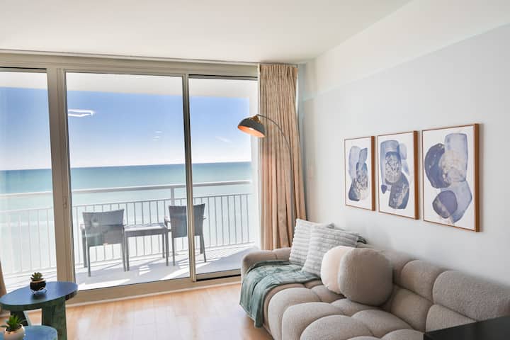 Las ventanas del piso al techo muestran vistas despejadas de las olas brillantes, llenando el espacio con luz natural y creando un ambiente sereno y relajante. La habitación está cuidadosamente amueblada con asientos lujosos y decoración contemporánea,