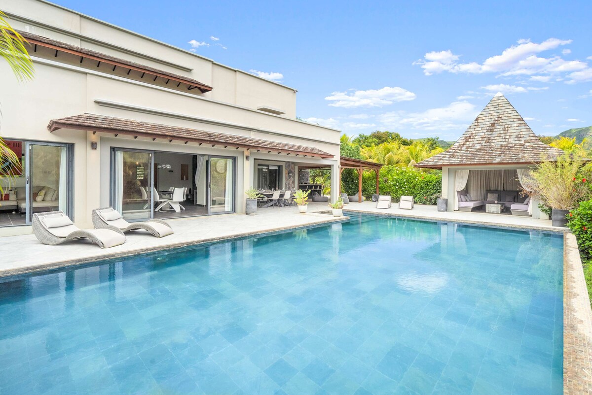 An infinity pool is positioned in the foreground, featuring clear blue water and sun loungers nearby. A gazebo with a thatched roof is visible in the background, surrounded by tropical greenery. The modern villa showcases large glass doors leading to a spacious patio.