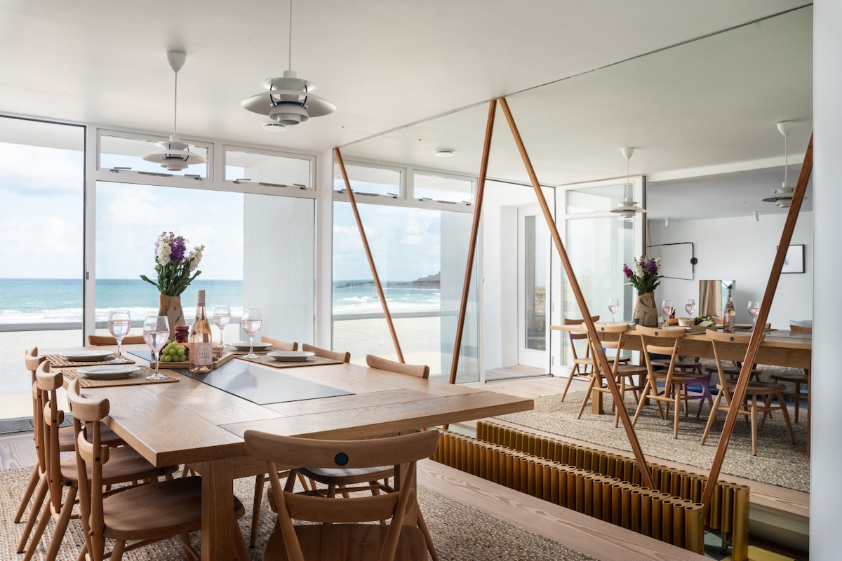 An open dining area features a large wooden table capable of seating eight, surrounded by stylish chairs. Mirrored walls reflect beach views and natural light, while a vase of fresh flowers adds a touch of color. The space connects to a private balcony through sliding glass doors.