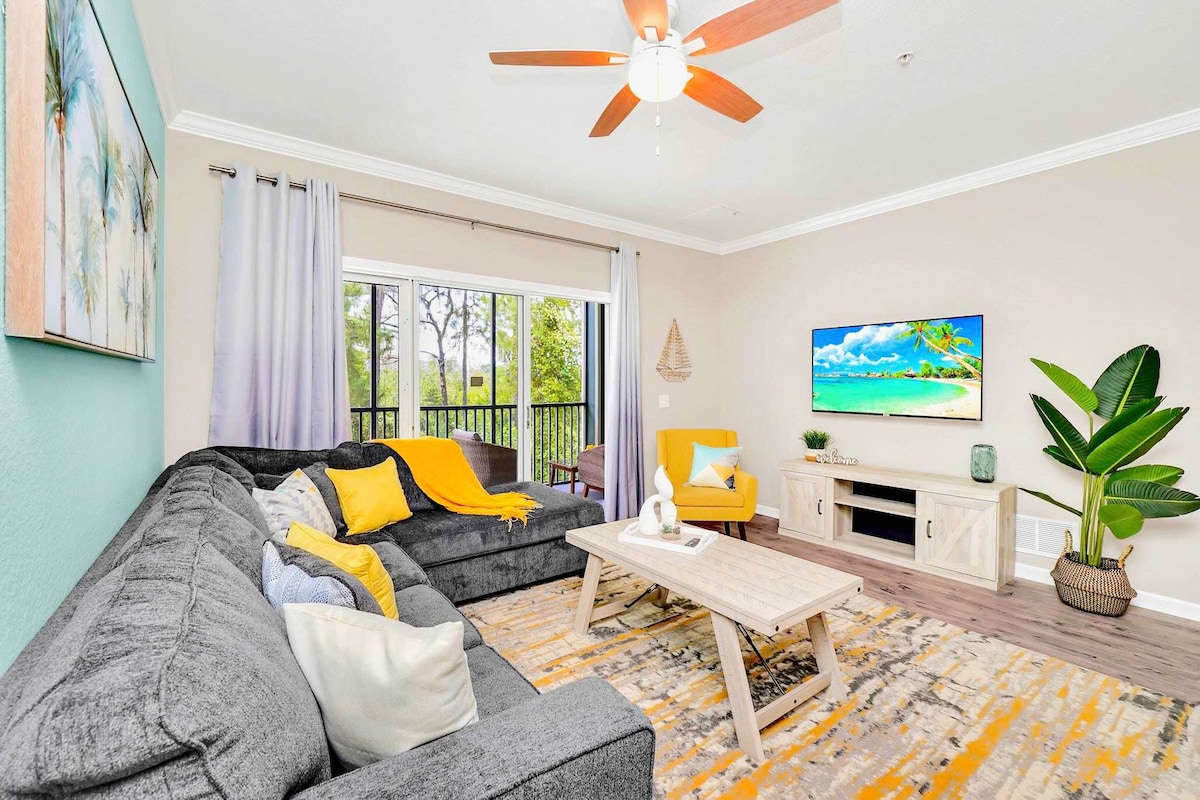 A modern living space is illuminated by natural light streaming through large glass doors. Comfortable seating is provided by a gray sofa and a bright yellow chair. A coffee table sits in the center, with decorative cushions and a vibrant area rug underfoot, complemented by a tropical view outside.