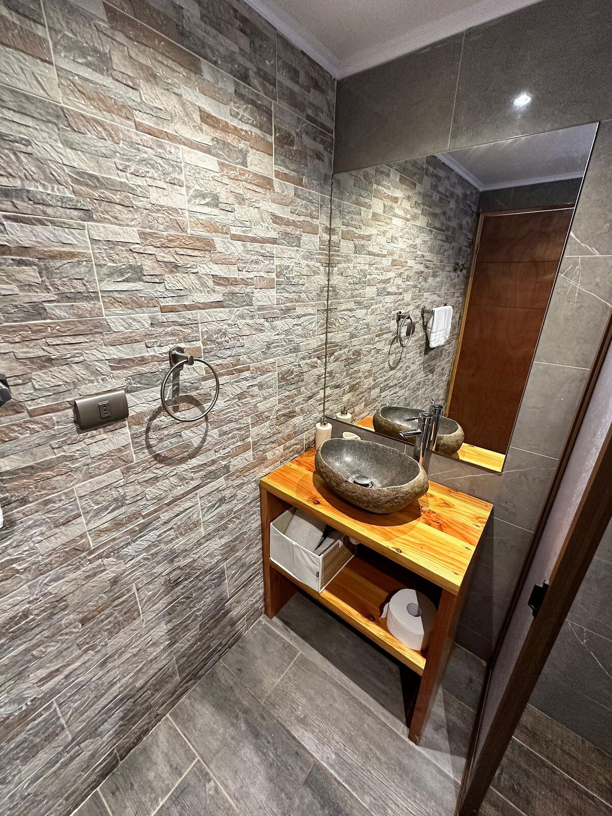 A modern bathroom features textured stone walls and a wooden vanity. A round, dark stone sink sits atop the natural wood surface, complemented by a large mirror that reflects the space. Towel rings and a dispenser are mounted nearby, providing necessary amenities.