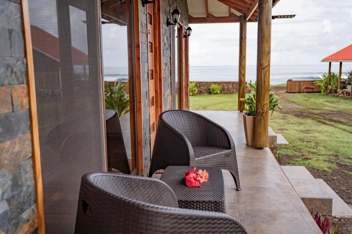 A covered patio features two woven chairs arranged beside a table. A vibrant flower rests on the table's surface, with a view of the ocean visible through large glass doors. Lush greenery surrounds the area, enhancing a sense of openness.