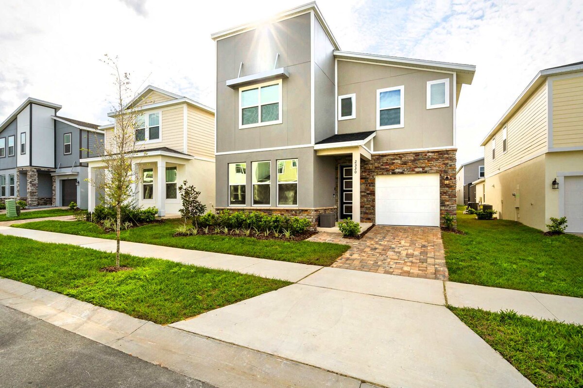 A modern two-story home is featured, showcasing a combination of stone and stucco exterior. The front is lined with well-manicured grass and small shrubs. A paved driveway leads to a garage, with a wide sidewalk providing easy access to the entrance.