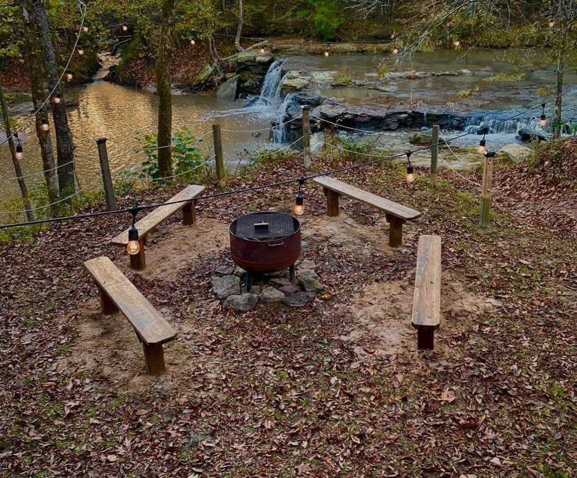 A circle of wooden benches surrounds a fire pit, situated on a sandy patch in the woods. Soft lighting from hanging lanterns creates a warm ambiance, while a waterfall can be seen in the background, alongside a serene body of water flowing gently.