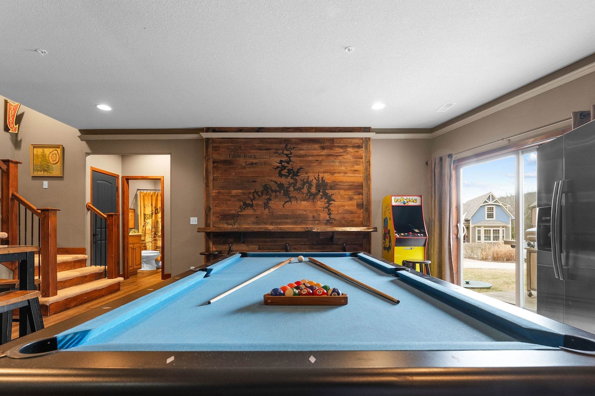 A game room is featured, showcasing a full-size pool table with a blue felt surface and a wooden triangle holding colorful balls. A wooden wall with a lake map adds visual interest, while arcade games are visible in the background along with doorways leading to other areas.