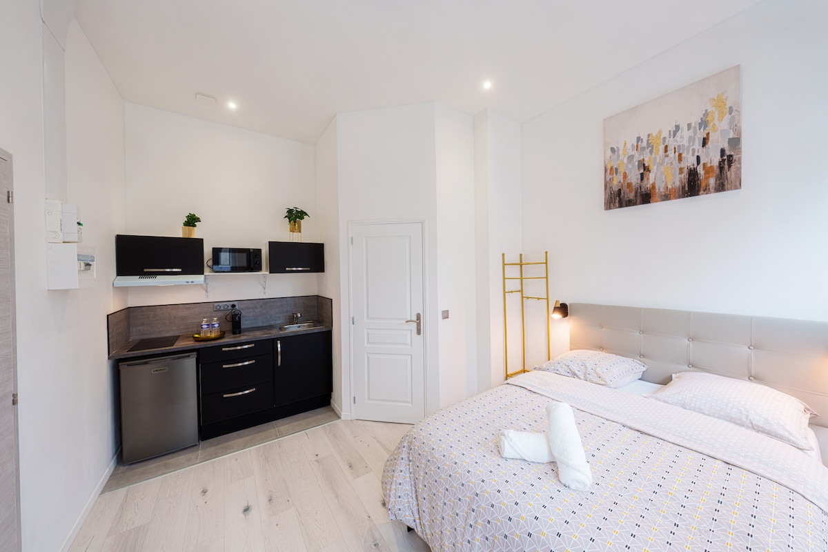 A modern studio space features a sleeping area with a spacious bed, quality linens, and bright natural light. The kitchenette is equipped with sleek black cabinetry, a refrigerator, microwave, and other essential appliances. A decorative piece hangs above the bed, contributing to the overall aesthetic.