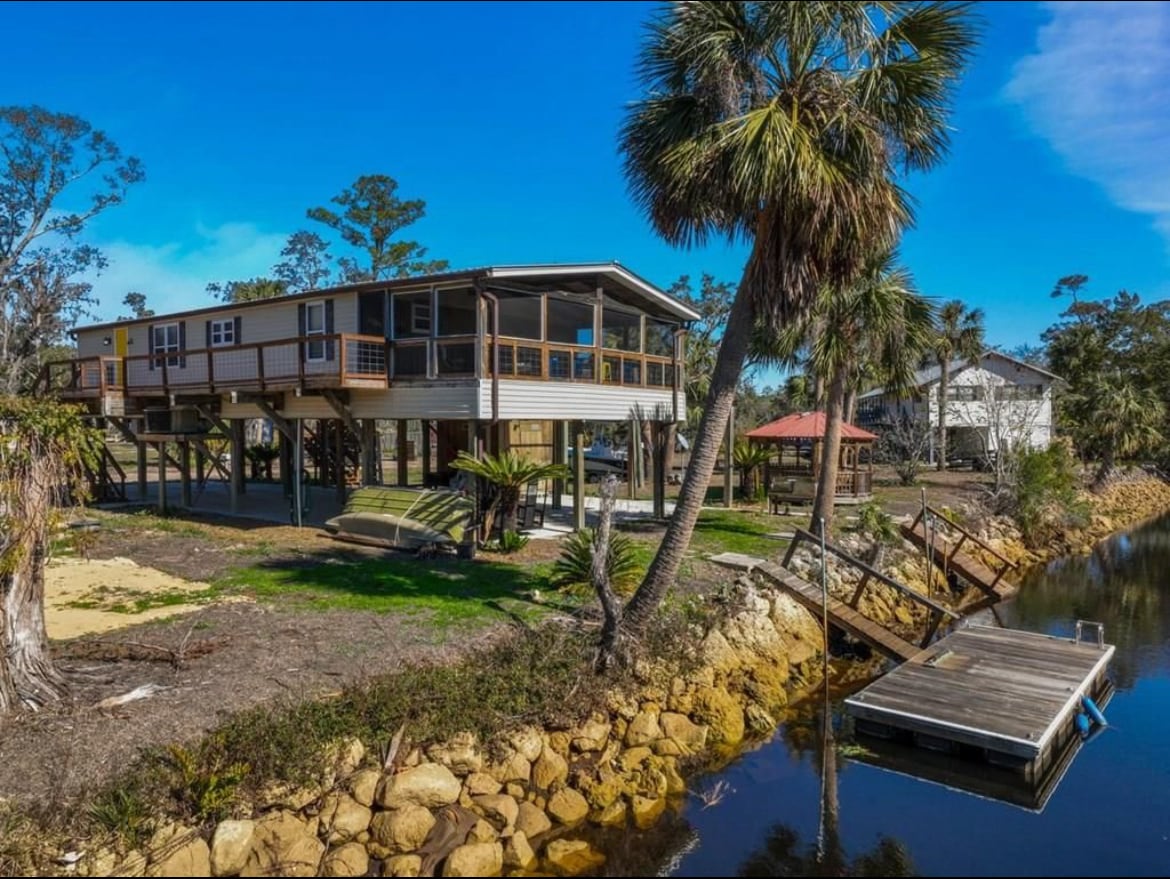 A raised vacation home is positioned near the water, surrounded by palm trees and greenery. A private dock extends into the water, providing access for boats. The area features open space with serene views of nature and a sunny blue sky.