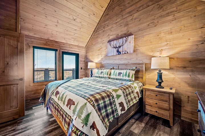 Со сводест дрвен таван, меко осветлување и постелнина инспирирана од природата, оваа спална соба зрачи со топлина и удобност. Преголемиот кревет е опкружен со две стилски ноќни шкафчиња, додека природните тонови на дрвената ламперија придонесуваат за удобниот естет на дрвената колиба