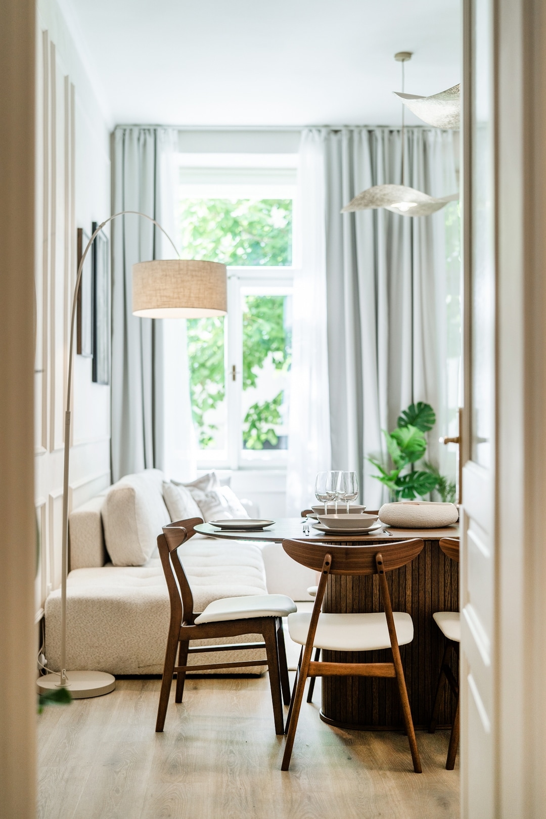 A modern living area features a light-colored sofa beside a round dining table with white chairs. Natural light flows through large windows adorned with sheer curtains, highlighting greenery placed nearby. Stylish pendant lights hang above the dining space, contributing to a cozy atmosphere.