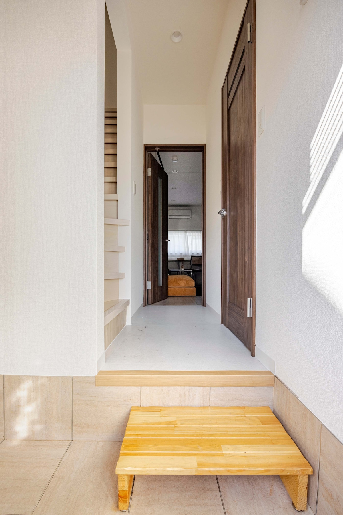 The entrance features a step up leading into a bright hallway, which is complemented by light coming through nearby windows. A small wooden step stool is positioned at the entrance, with a glimpse of staircase steps visible on the left.