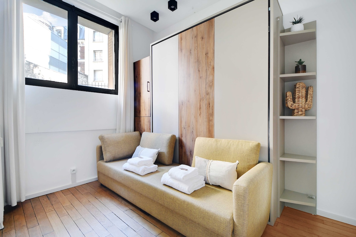 A compact living space features a sleek, modern sofa bed adorned with neatly arranged bath towels. The space is enhanced by a tall shelving unit displaying a decorative cactus and minimalistic decor. Natural light streams in through the large windows, accentuating the warm wooden floor.