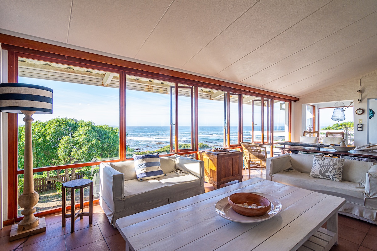 An airy living area features large windows that provide panoramic ocean views. Comfortable white couches and a wooden coffee table are arranged thoughtfully, allowing for relaxation. Natural light floods the space, enhancing the open atmosphere that connects to a dining area set for gatherings.