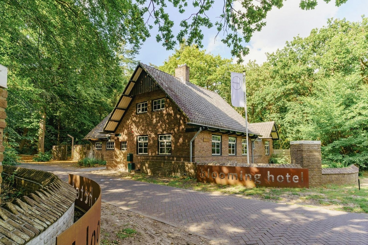The exterior of a charming villa is nestled within lush trees, featuring a sloped roof and numerous windows that provide natural light. The entrance is marked by a sign displaying 'blooming hotel,' set against a backdrop of serene forest foliage.