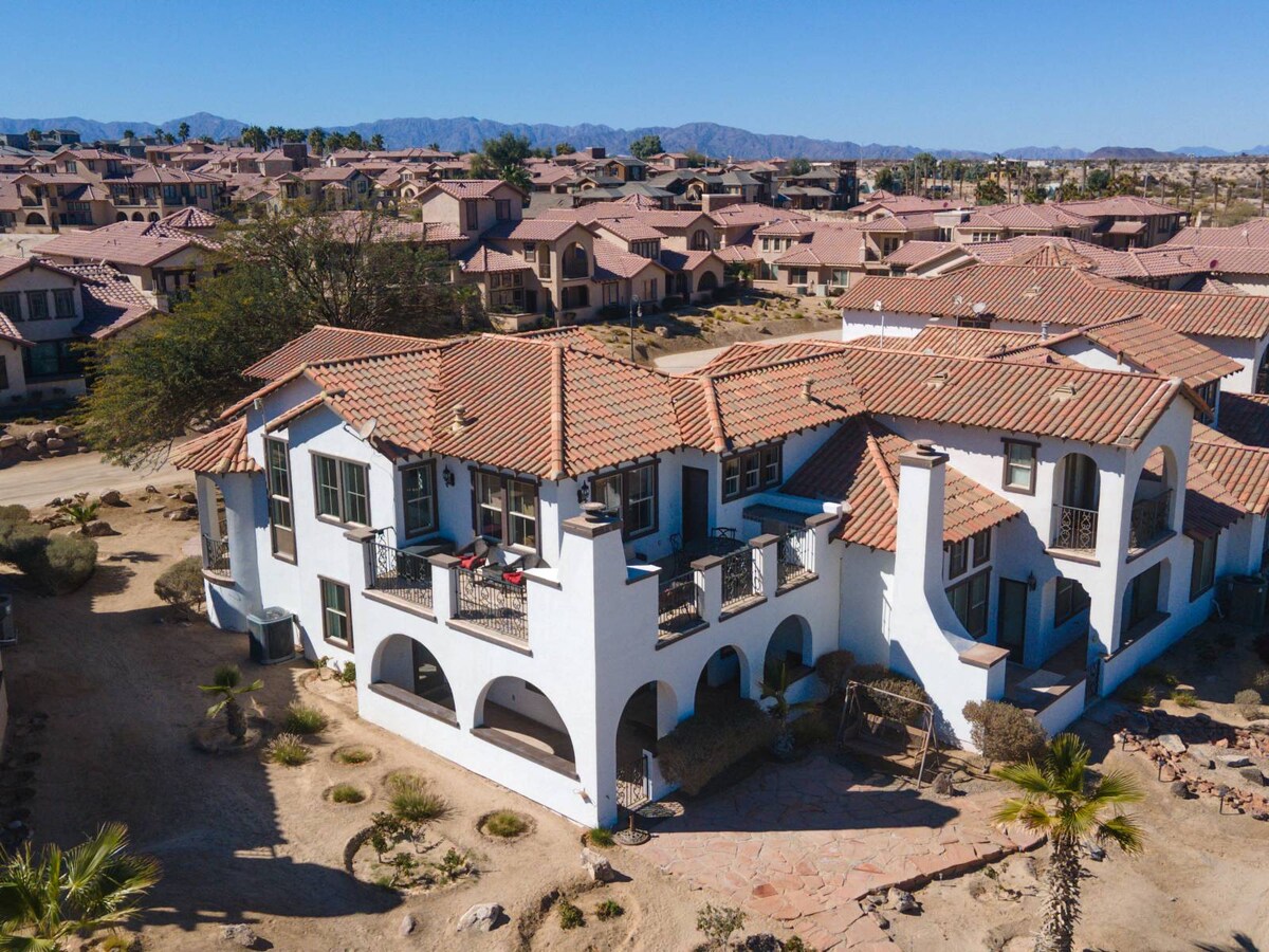 An aerial view of the two-story condo showcases its distinctive white exterior and terracotta roof tiles. The surrounding landscape features desert vegetation and neighboring homes, complemented by the foothills in the background. Outdoor seating is visible on the balcony, providing an inviting space for relaxation.