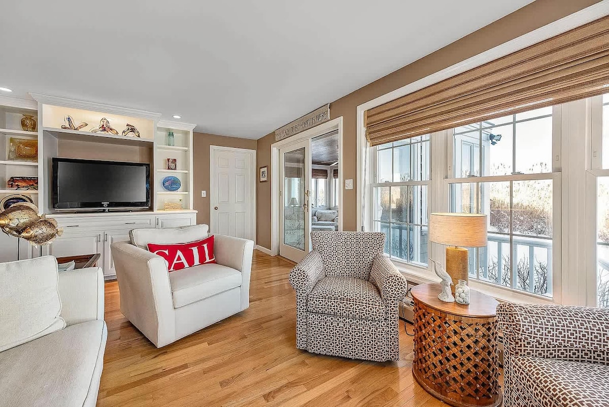 A cozy living area features a comfortable sofa and two armchairs, inviting relaxation. A flat-screen TV is mounted on a white entertainment unit. Large windows offer ample natural light and views of the outdoors, while a stylish side table with a lamp adds a touch of warmth.