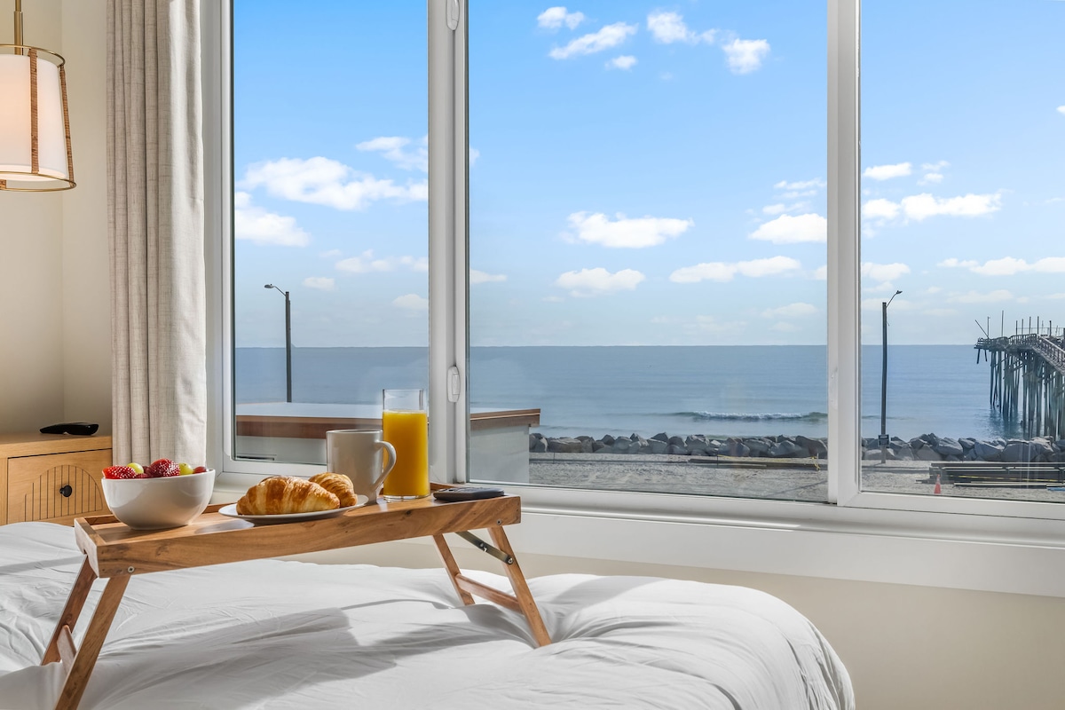 A breakfast tray with fruit, juice, and a croissant is placed on the bed, with a view of the ocean visible through large windows. The serene water reflects the clear blue sky, offering a peaceful coastal ambiance.