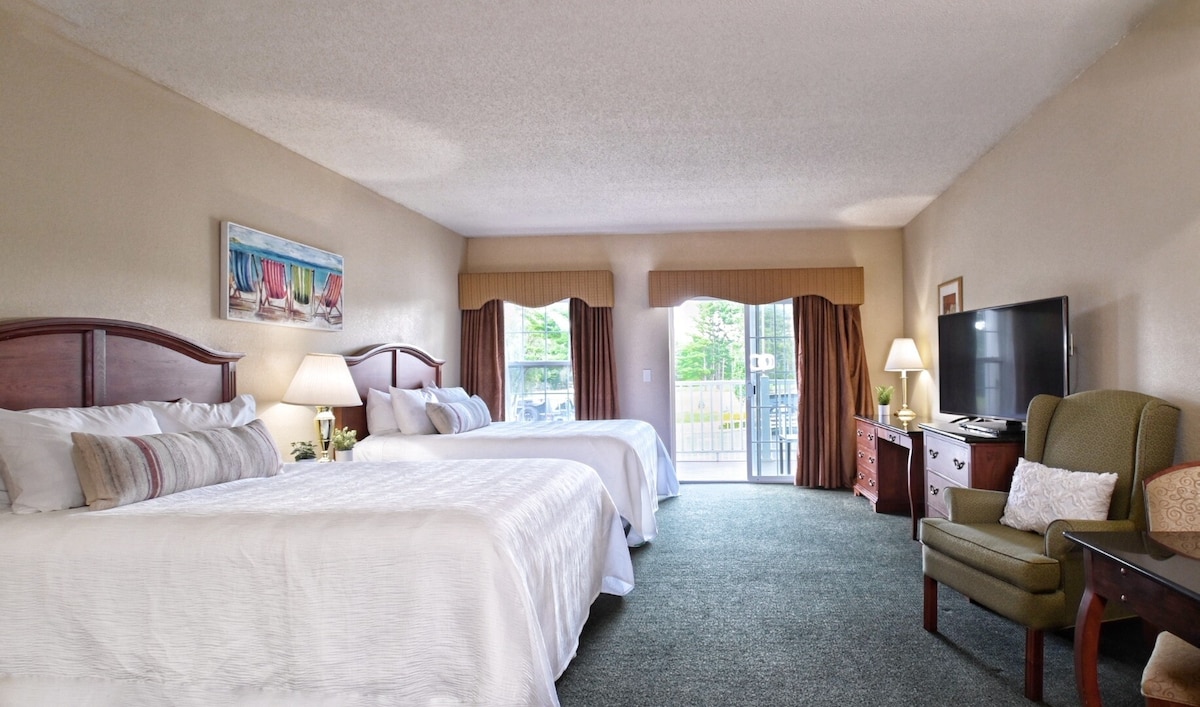 A spacious hotel room features two large beds dressed in white linens. Natural light enters through sliding glass doors, flanked by curtains. A large flat-screen TV is positioned against the wall, and a comfortable armchair is positioned nearby, enhancing the room's functional layout.