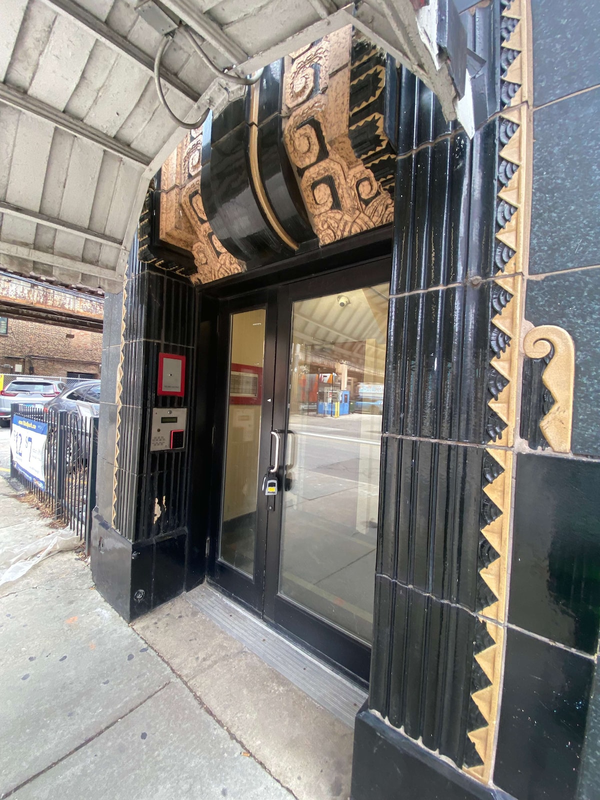 The main entrance features a sleek black door surrounded by detailed decorative elements in gold and cream tones. The entryway is partially covered by an awning, providing shelter from the weather. A minimalist mailbox can be seen to the left of the door.