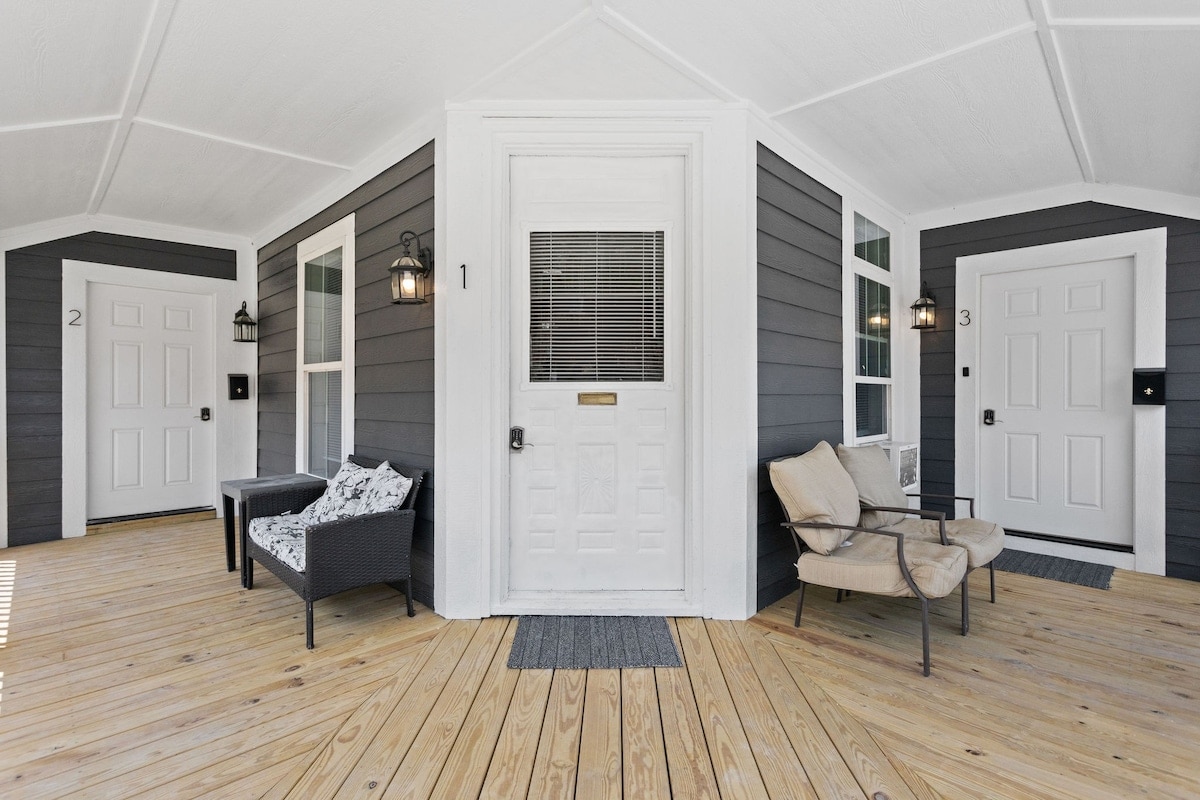 A welcoming entrance features a spacious wooden deck with two seating areas, showcasing a pair of chairs and a small table. Three doors lead into the property, framed by dark grey siding and accentuated by elegant lanterns for added illumination.
