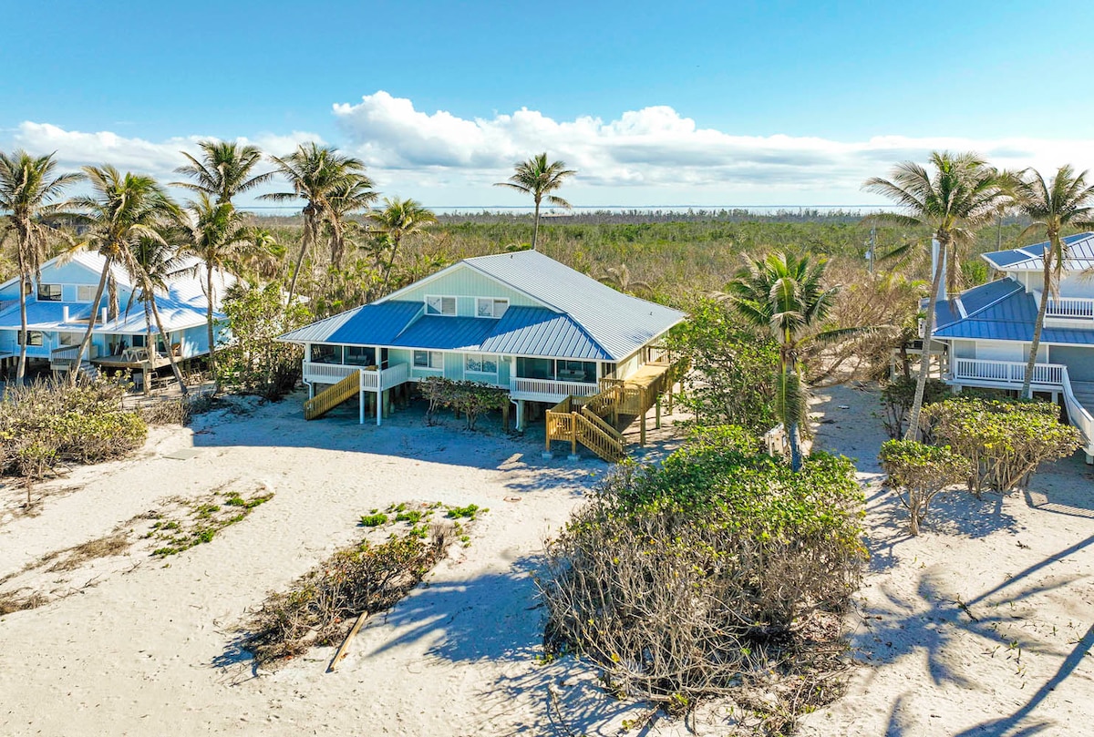 A spacious beach-front townhome is nestled among tall palm trees, with a sandy area leading directly to the shoreline. The building features a blue metal roof and a wooden deck, offering a welcoming view of the surrounding landscape under a clear blue sky.