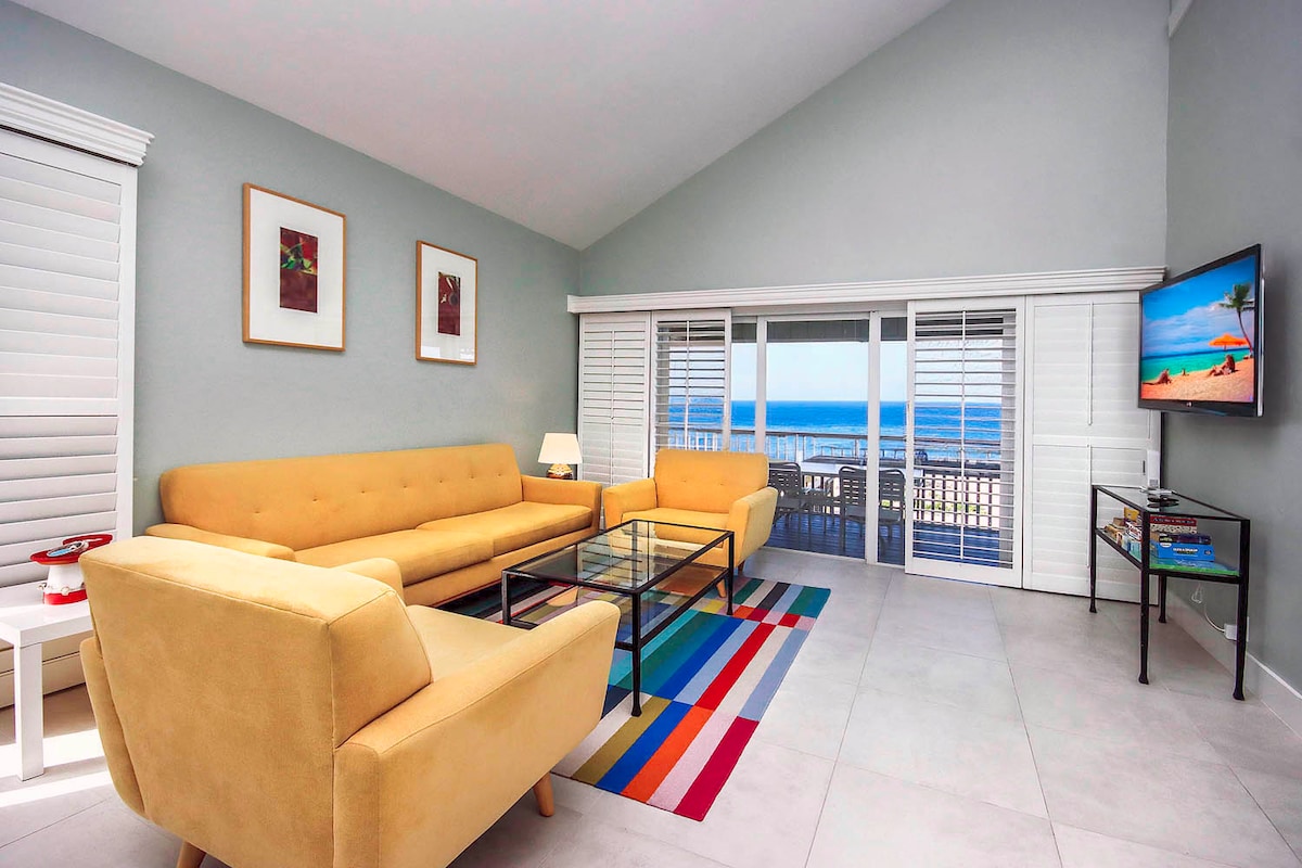 A bright and airy living area features a large glass coffee table surrounded by a yellow sectional sofa and two matching chairs. Natural light floods in through sliding glass doors, revealing views of the Gulf outside. A flat-screen TV is mounted on a wall, alongside a colorful area rug.