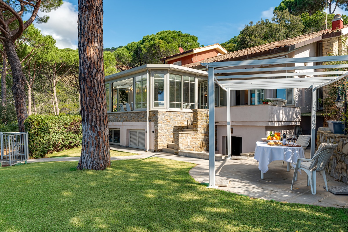 An outdoor dining area is showcased under a white pergola, featuring a table set with fresh fruit. A spacious garden is framed by trees, with a stone facade visible on the villa. Large windows offer a view of the surrounding landscape.