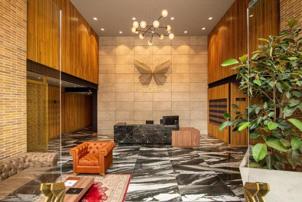 A modern lobby area is highlighted by elegant marble flooring and warm wooden accents. A decorative butterfly emblem is positioned on the wall behind the reception desk. Cozy seating is provided, including a brown leather couch, creating an inviting entry space.