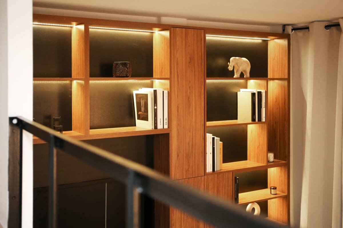 A stylish wooden shelving unit is illuminated by soft, warm lighting. Various decorative items, including a small white elephant and books, are displayed on the shelves. The unit provides a modern and organized look within the apartment's living space.