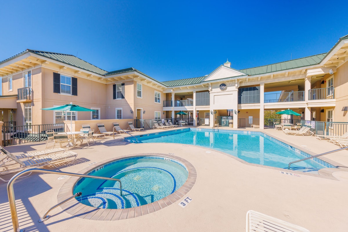 The outdoor pool area features a clear, inviting pool surrounded by lounge chairs and umbrellas. A hot tub is positioned nearby, with a gentle curvature. The buildings framing the pool showcase a light color, reflecting a welcoming environment under a bright blue sky.