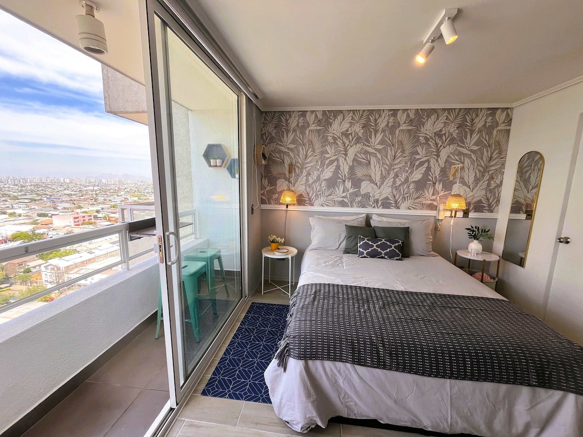A well-lit studio features a spacious bed dressed in neutral linens, complemented by patterned wallpaper. A small table and chairs are positioned near floor-to-ceiling glass doors, which open to a balcony with expansive city views. Decorative elements include wall sconces and a vase of flowers.
