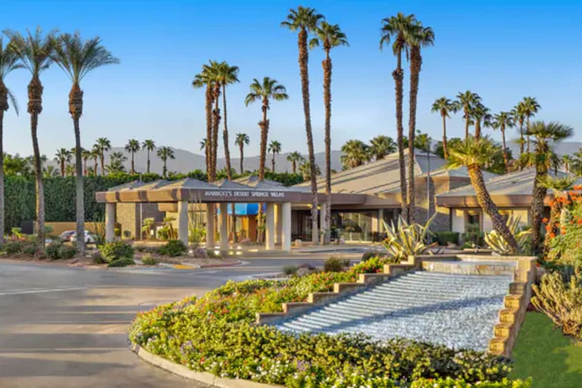 The entrance to Marriott’s Desert Springs Villas is lined with palm trees and vibrant landscaping. A water feature flows gently in front, leading to the main entrance, which showcases a welcoming design set against a clear blue sky.