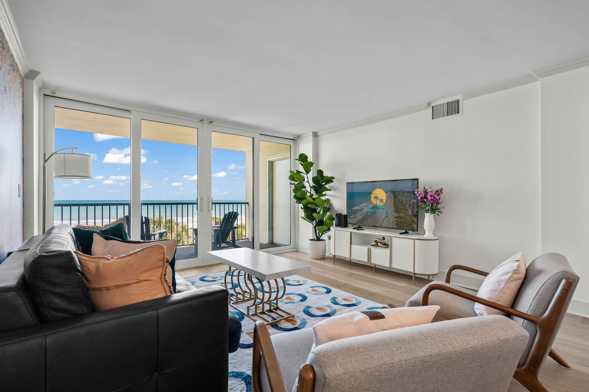 A bright family room features large glass doors that provide views of the ocean. Comfortable seating includes a black sofa and two wooden chairs. A coffee table is centered on a vibrant area rug, while a green plant adds a touch of nature to the space.