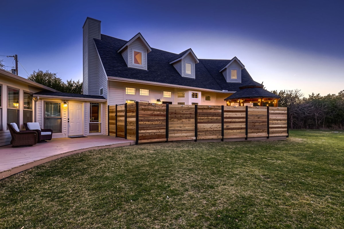 The exterior of the home is framed by a spacious grassy yard and a fenced patio area. Soft outdoor lighting highlights the structure, which features a combination of gabled roofs and multiple windows. Relaxation areas are visible, including seating positioned near the entrance.