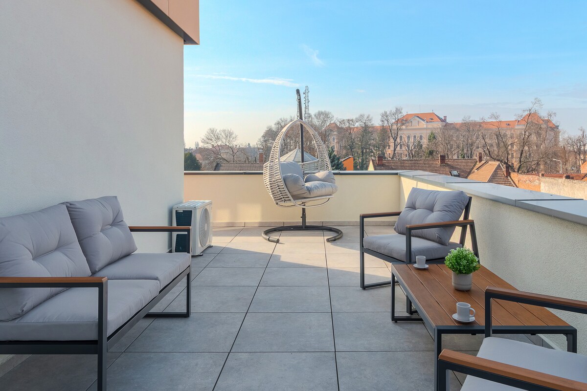 A spacious private terrace is presented with modern outdoor furniture, including a hanging chair and two cushioned armchairs surrounding a coffee table. Clear skies and nearby greenery complement the inviting space, perfect for relaxation and enjoying panoramic views of the city.