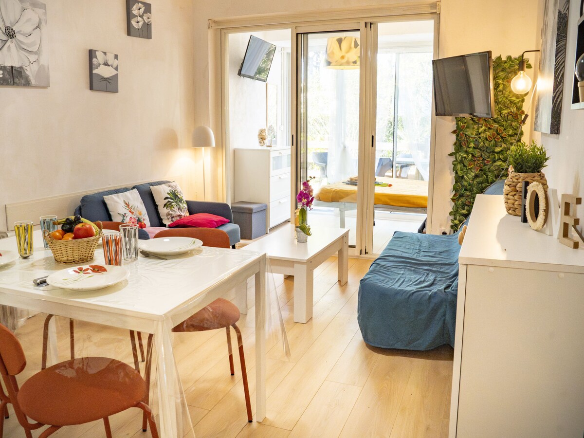 The living area is furnished with a blue sofa and a dining table set for four. A flat-screen TV is mounted on the wall. Large glass doors offer access to an outdoor terrace, while natural light fills the room, creating a bright and airy ambiance.